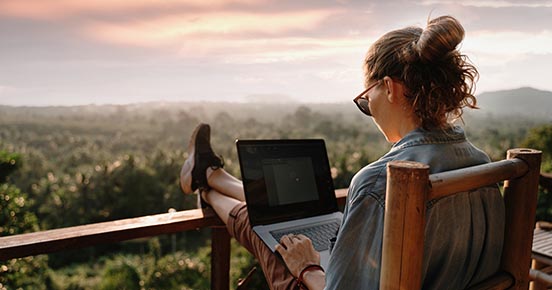 Imagine cu o tânără programatoare admirând un peisaj deosebit de pe balcon, cu laptopul în brațe, ilustrând flexibilitatea și libertatea pe care le oferă învățarea online prin cursurile de programare de la Coders Lab.
